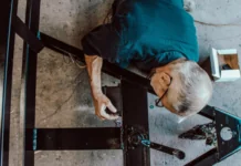 SVP-Initiative: «Länger arbeiten? Das wäre realistisch» Vogelperspektive auf einen älteren Mann mit grauen Haaren, der eine Brille und ein blaues Hemd trägt. Er bearbeitet elektrische oder mechanische Komponenten an einer schwarzen Metallstruktur, die auf dem Boden liegt. In seiner Nähe sind Werkzeuge und eine kleine weiße Schachtel zu sehen.