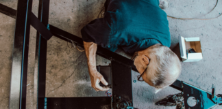 SVP-Initiative: «Länger arbeiten? Das wäre realistisch» Vogelperspektive auf einen älteren Mann mit grauen Haaren, der eine Brille und ein blaues Hemd trägt. Er bearbeitet elektrische oder mechanische Komponenten an einer schwarzen Metallstruktur, die auf dem Boden liegt. In seiner Nähe sind Werkzeuge und eine kleine weiße Schachtel zu sehen.