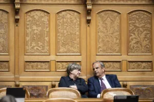 Deux personnes en tenue formelle sont assises côte à côte dans un cadre officiel. Elles sont positionnées devant un mur orné de boiseries dorées richement décorées, caractérisé par des panneaux sculptés présentant des motifs floraux et figuratifs élaborés. Les chaises sur lesquelles elles sont assises sont de style classique avec dossiers en rotin. La femme à gauche, vêtue d'une veste noire, regarde vers la droite, tandis que l'homme à droite, portant un costume bleu marine avec une cravate, se tourne légèrement vers elle. L'arrière-plan somptueux suggère un lieu prestigieux tel qu'une institution gouvernementale, un tribunal ou une organisation importante.