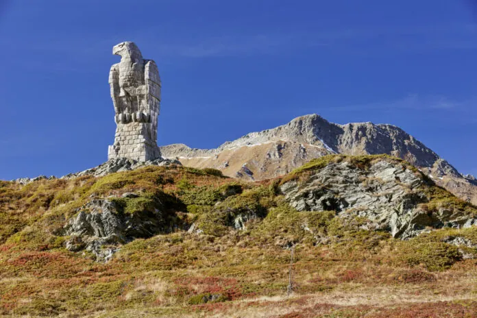 Eine grosse, aus hellem Stein gemauerte Adlerstatue thront auf einem gemauerten Sockel auf einer felsigen Bergkuppe. Der Adler hat angelegte Flügel und blickt zur Seite. Im Hintergrund erstreckt sich eine karge, felsige Berglandschaft mit vereinzelten Schneeresten unter strahlend blauem Himmel. Der Vordergrund zeigt eine hügelige alpine Wiese mit herbstlich rot-braun und grün gefärbter Vegetation sowie freiliegenden Felsformationen.