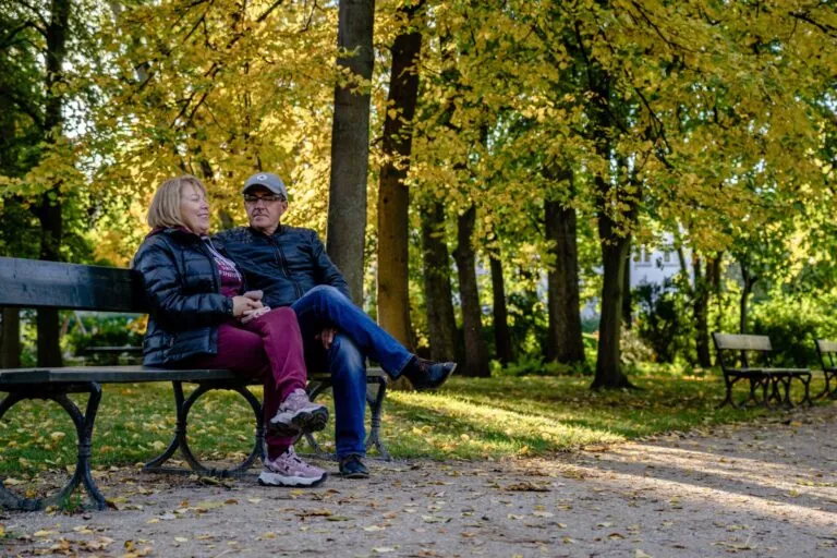 Zwei Personen mittleren Alters sitzen auf einer Bank in einem Park und schauen sich lächelnd an. Sie sind herbstlich gekleidet mit Jacken. Der Mann trägt eine Mütze und eine dunkelblaue Jacke, die Frau eine marineblaue Jacke über einer bordeauxfarbenen Hose. Hinter ihnen erstreckt sich eine Allee mit hohen Bäumen, deren Laub in gelben und grünen Tönen leuchtet. Der Boden ist mit Herbstblättern bedeckt. Im Hintergrund sind weitere Parkbänke und Gebäude sichtbar. Die Szene vermittelt einen Eindruck von Ruhe und gemeinsamer Zeit in der Natur.