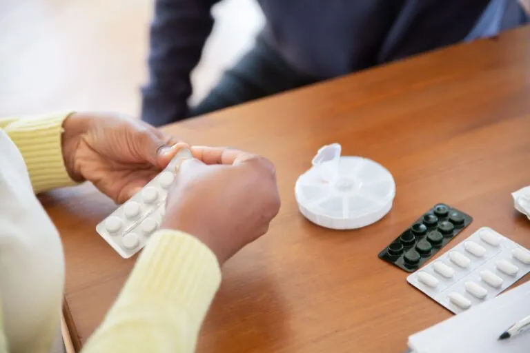 Nahaufnahme von zwei Händen, die eine Tablettenblisterpackung öffnen, an einem Holztisch mit einer runden Pillendose sowie weiteren Blisterpackungen mit weissen und dunklen Tabletten.